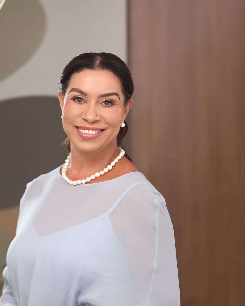 Marta Benfica, de blusa azul-clara e colar de pérolas, em pé no escritório, sorrindo.
